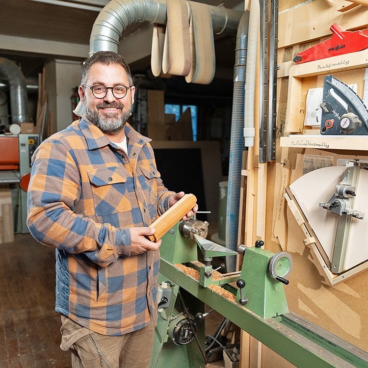 Holzhandwerk Kohler Ein Mann steht vor einer Drechselbank und hält ein Stück Holz.